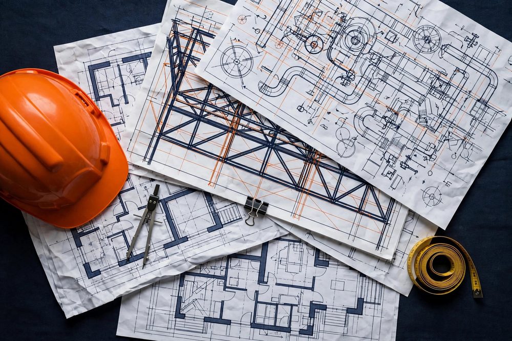 Architectural, structural and mechanical drawings layered on a construction table with a hard hat and measuring tape